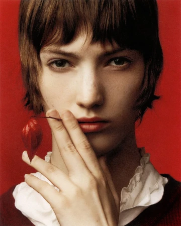 Portrait of a brunette female model with her hand near her face, holding a red chinese lantern plant against a red studio backdrop.