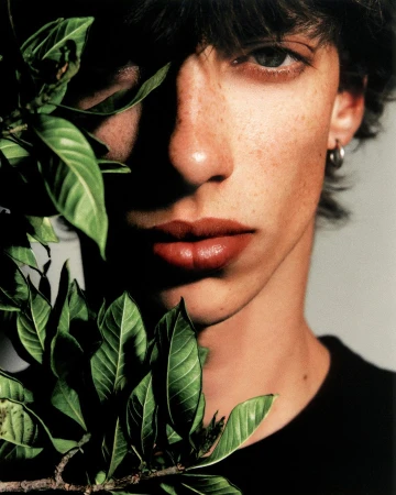 Portrait of a young adult male with freckles and dark hair, posing indoors with green leaves partially covering his face.