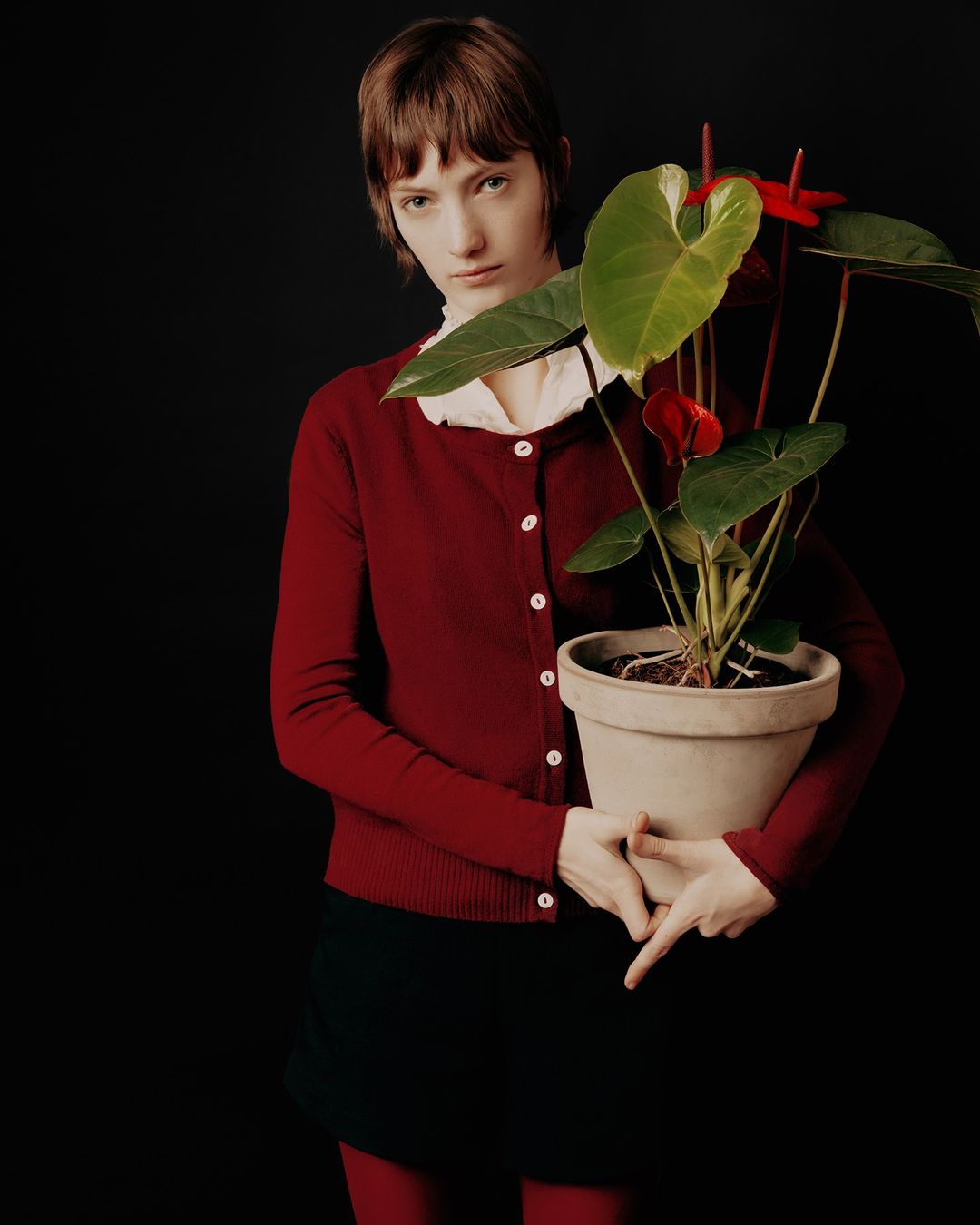 A young adult woman with bangs, wearing a red cardigan, holds a potted anthurium plant against a solid black backdrop in a studio shot.