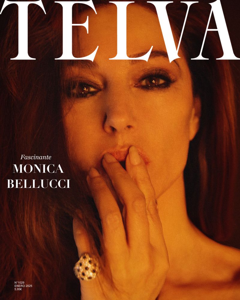 Monica Bellucci gazes into the camera on a magazine cover. Fingers touch her lips, showcasing a large ring, bathed in warm, studio lighting.