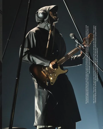 Man in hooded coat plays electric guitar on stage with microphone. Dark, moody scene with metal poles and audio equipment.