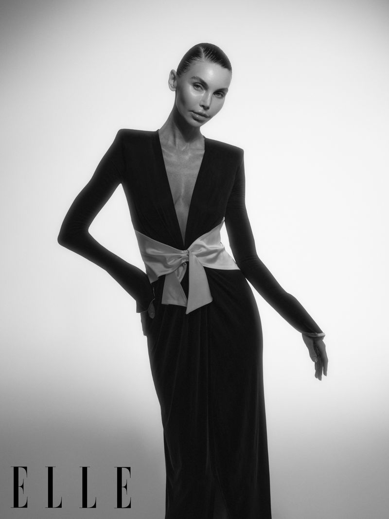 Black-and-white fashion portrait of a woman in an elegant dress with a bow, posing against a gradient white backdrop in a studio.