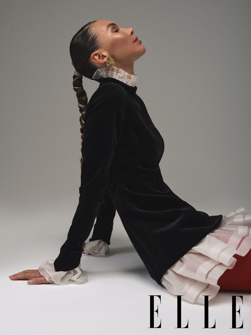 Portrait of a woman in a black velvet dress with a ruffled collar and long braided hair, posing against a grey background.