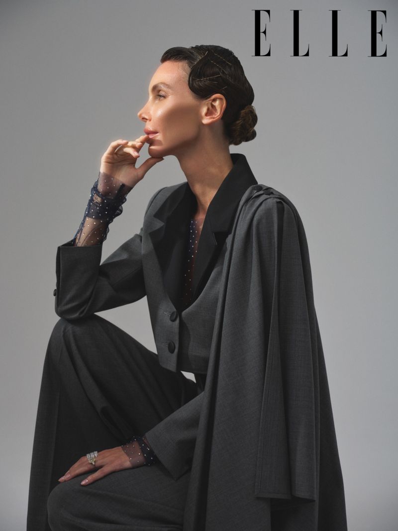 Elegant woman in dark suit poses indoors in front of grey background. She is wearing sheer dotted sleeves and has her hand to her face.