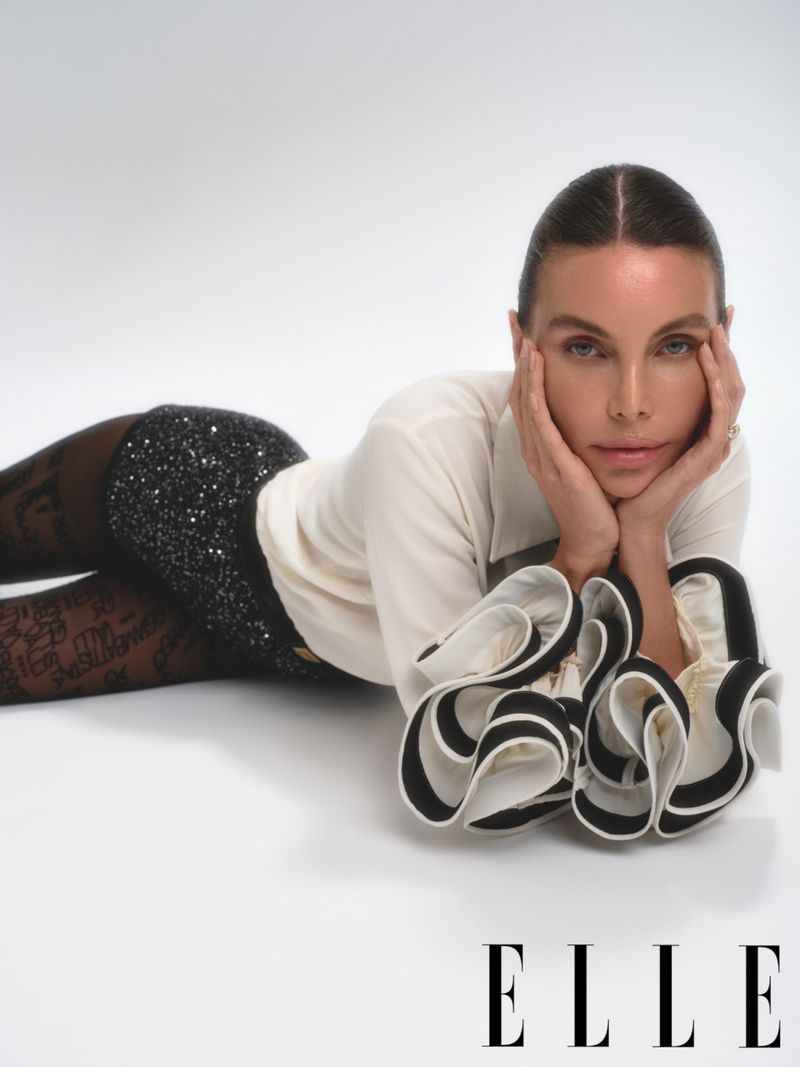 Adult woman with a neutral expression poses on a white surface, wearing a long-sleeve shirt, a sequined skirt, and patterned tights.