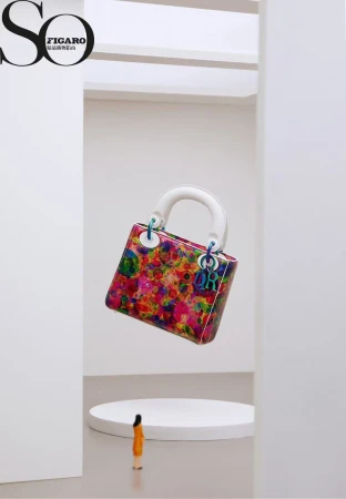 Colorful Dior handbag on a white pedestal, juxtaposed with a tiny model in an expansive white studio setting for a fashion editorial.