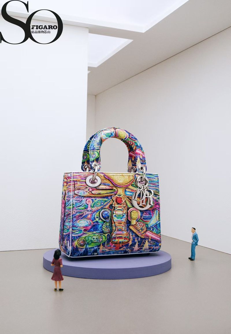 A large, colorful Dior Lady handbag stands on a purple platform between two miniature human figures in a clean, white exhibition space.