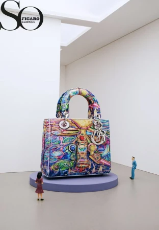 A large, colorful Dior Lady handbag stands on a purple platform between two miniature human figures in a clean, white exhibition space.
