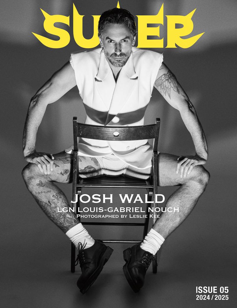 Josh Wald sits on a chair with his legs spread, showcasing tattoos, wearing a white vest and black dress shoes in a studio setting.