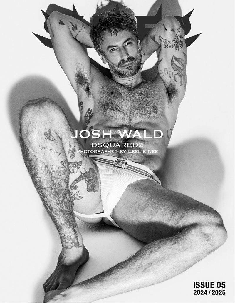 Black and white studio portrait of a graying male model lying on his side with visible tattoos and wearing a white jockstrap.