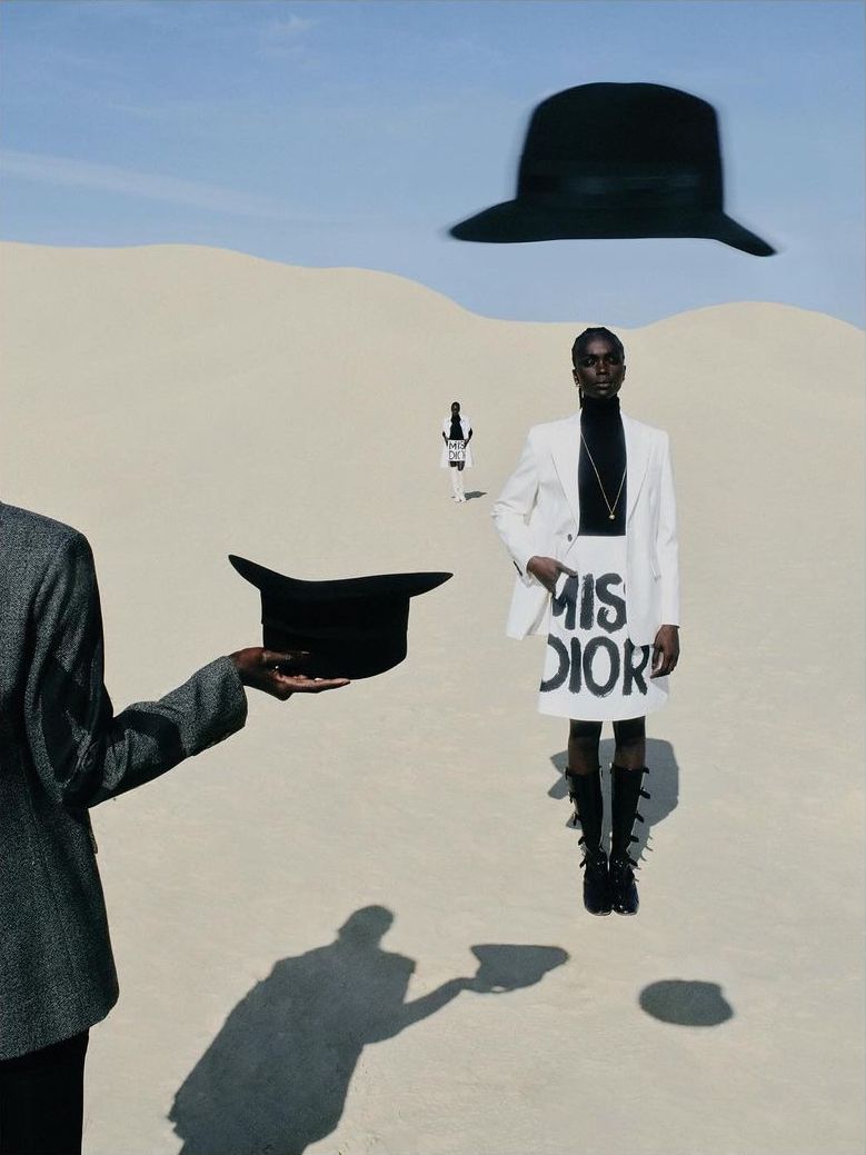 Fashion editorial shot of a Black female model in a white skirt suit in a desert landscape with blue sky and hats, with surreal elements.