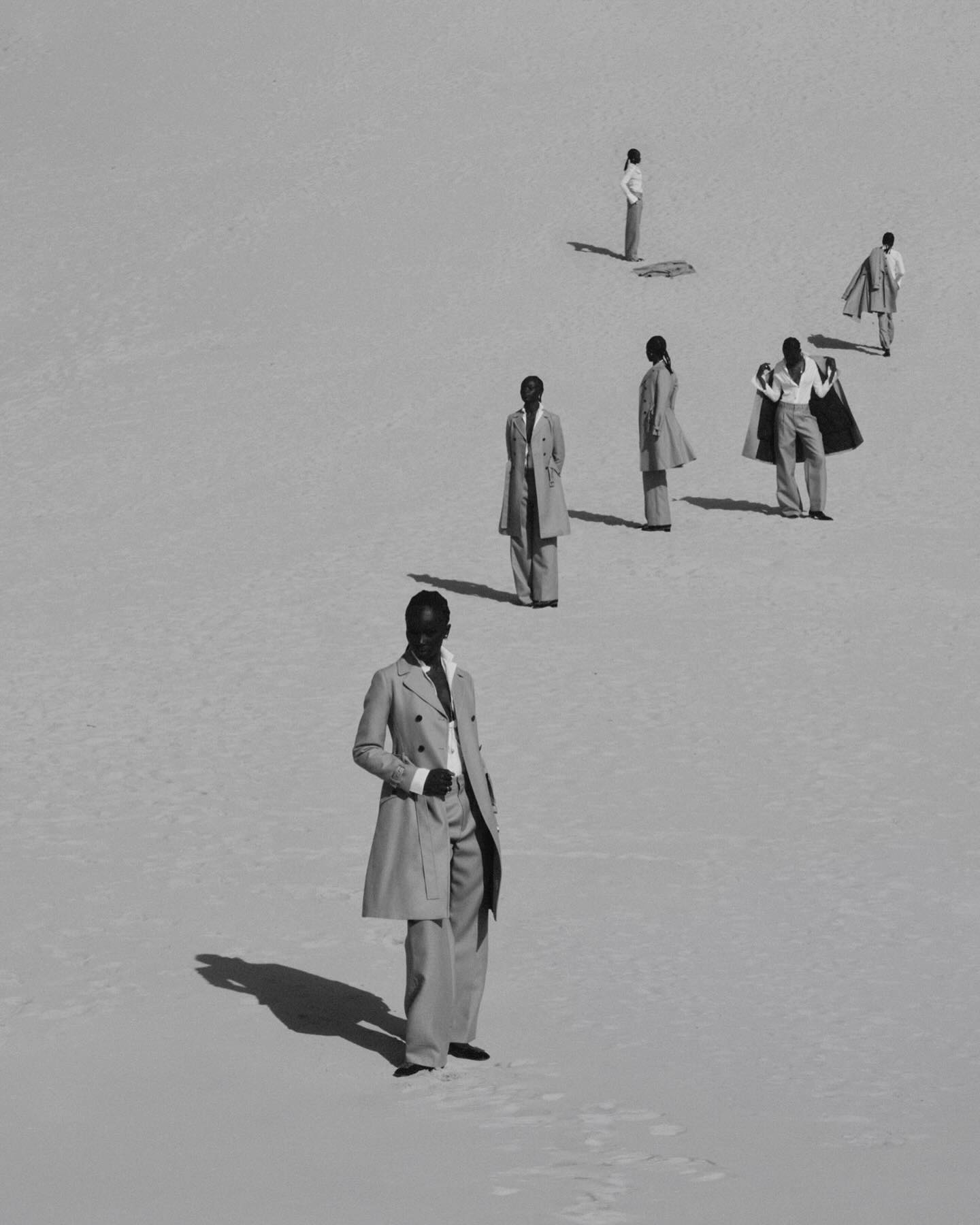 Fashion editorial shot in black and white: A group of Black models stand on a bright sandy landscape, wearing neutral-tone vintage suits.