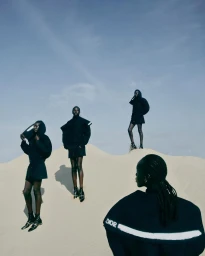 Four Black models in black hooded Dior outfits posing across sandy dunes under an overcast sky.