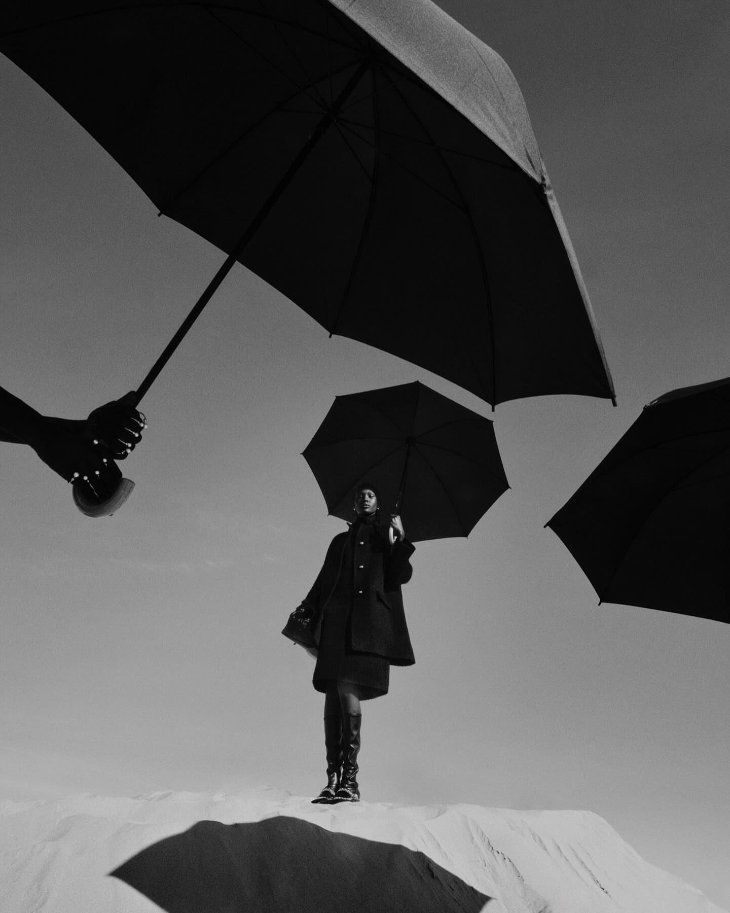 Monochrome outdoor fashion shot featuring a woman standing on a sandy surface, surrounded by dark umbrellas held by hands.