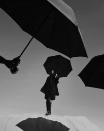 Model standing beneath multiple large black umbrellas held against a bright sky, wearing a dark coat and boots.