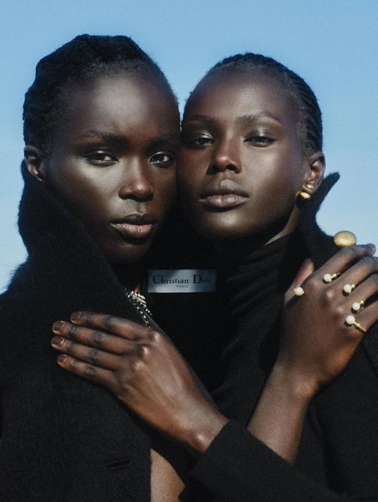 Two black female fashion models in black jackets posing closely in front of a blue sky, displaying gold jewelry and featuring Christian Dior branding.