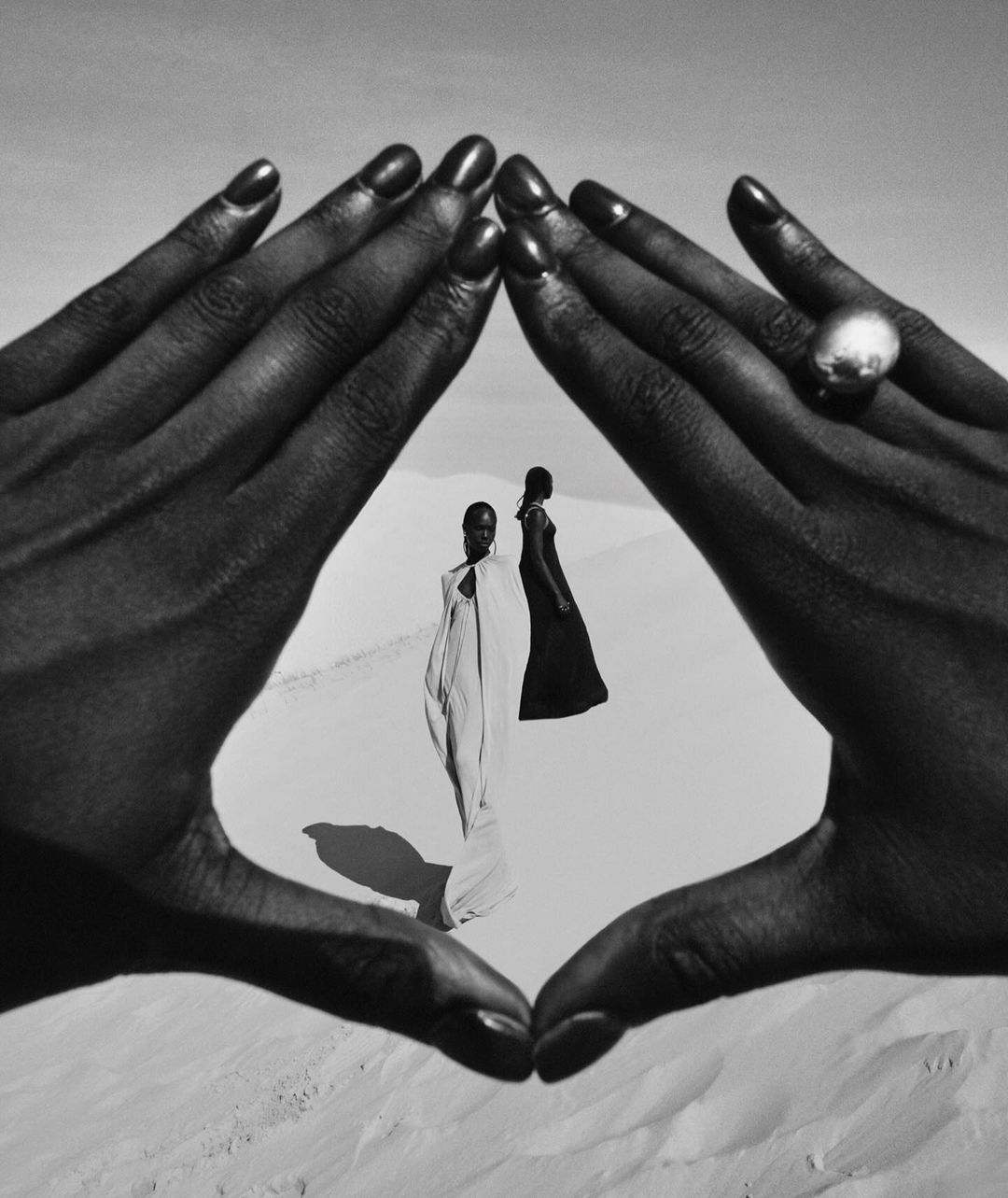 Black and white fashion portrait of two dark-skinned female models in draped gowns, framed by dark-skinned hands with a ring on sand.