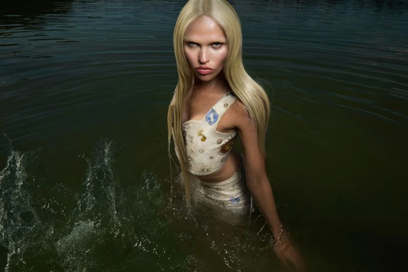 Adult woman with blonde hair, wearing a two-piece embellished garment, wading in dark water with splashes, portrait orientation.