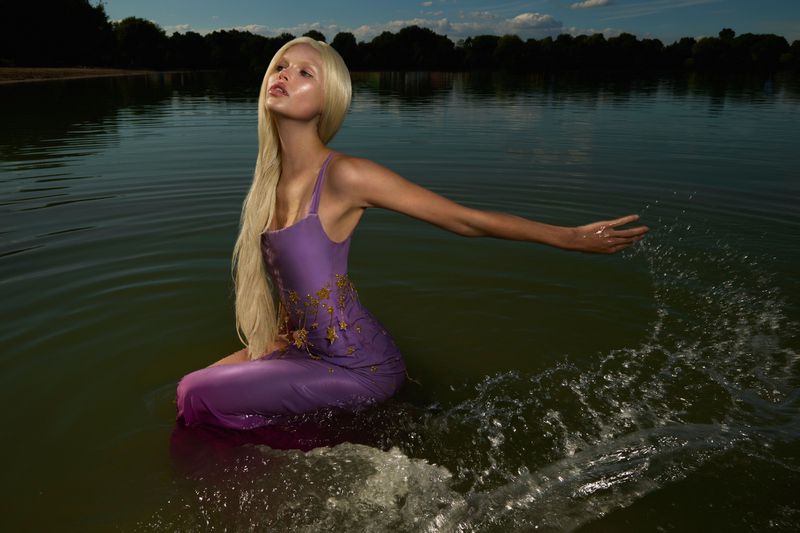 Woman with long blonde hair in lilac dress sitting in a lake with hand extended, creating water splashes, with trees and blue sky in background.