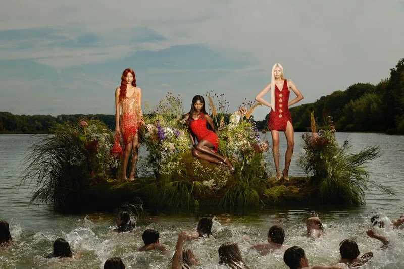 Three fashion models in red dresses pose on a floating floral platform on a lake with swimmers; trees and sky are in the background.