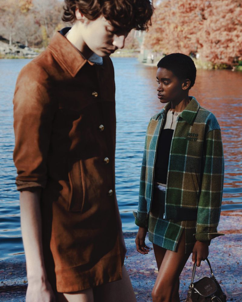 Two people pose outdoors near water; one in a brown jacket, the other in plaid with a purse. Fashion editorial shoot in nature.