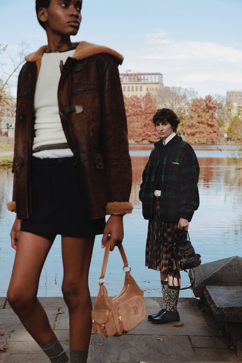 Two women modeling fall fashion in a park setting, one with a brown shearling coat and handbag, the other in a kilt with a green plaid jacket.