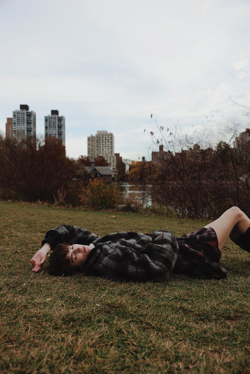 Model lying on a grassy lawn in an urban park wearing a dark plaid jacket and pleated skirt with buildings in the background.
