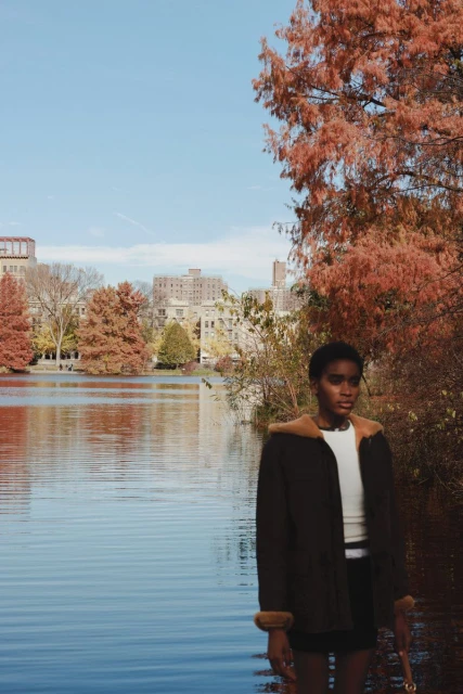 A stylish woman in a brown coat stands near a lake surrounded by fall foliage and city buildings in the background, on a bright day.