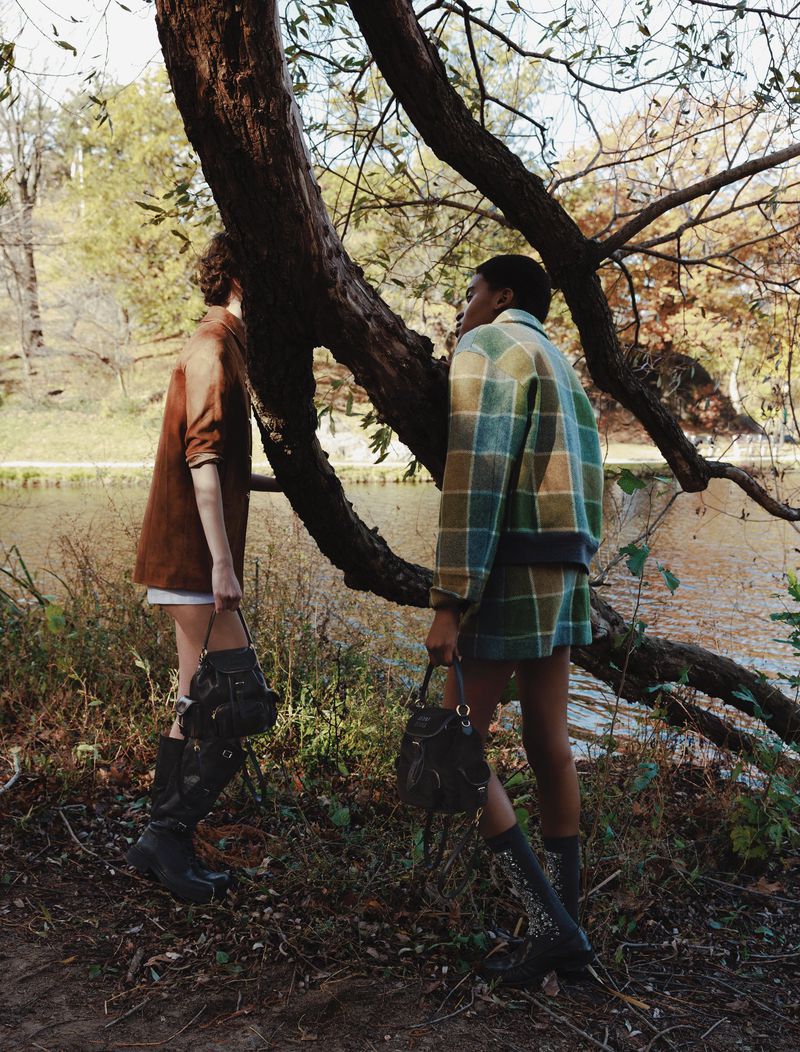 Two women in fall fashion stand near a tree and lake, wearing plaid and brown outfits with knee-high boots and matching mini-bags.