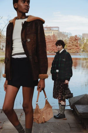 Model in a brown shearling coat holding a tan suede bag with a second model in plaid standing near a lake in autumn.