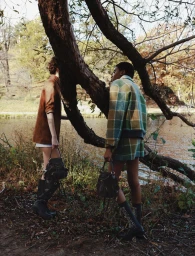 Two models posing in autumn outerwear and holding black designer handbags beside a tree trunk along a sunlit lakeside.