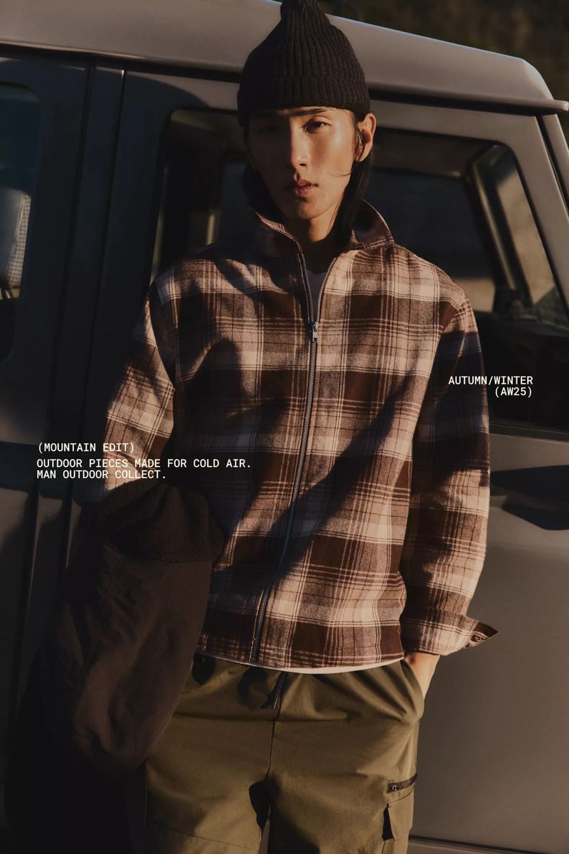 Male model poses outdoors in plaid jacket with a beanie hat, against a car and greenery, showcasing Autumn/Winter fashion.