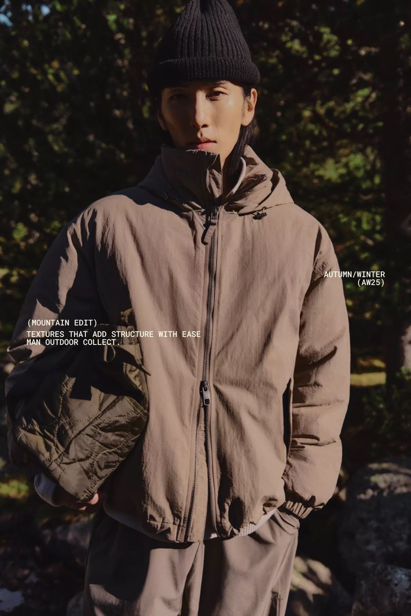Asian man wearing a dark beanie and khaki puffer jacket in outdoor setting with forest background, autumn/winter (AW25) ad campaign.