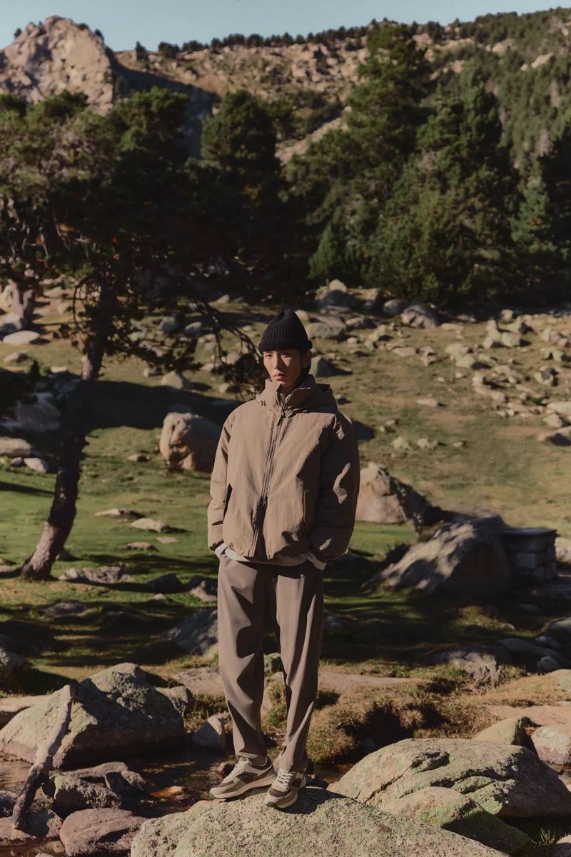 An Asian man in a brown hooded jacket and trousers standing on rocks outdoors with a mountain backdrop, capturing fashion and nature.