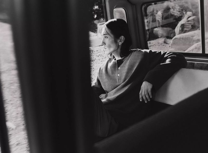 Black-and-white photo of a young adult leaning against a window inside a vehicle, gazing out at the outdoor landscape.