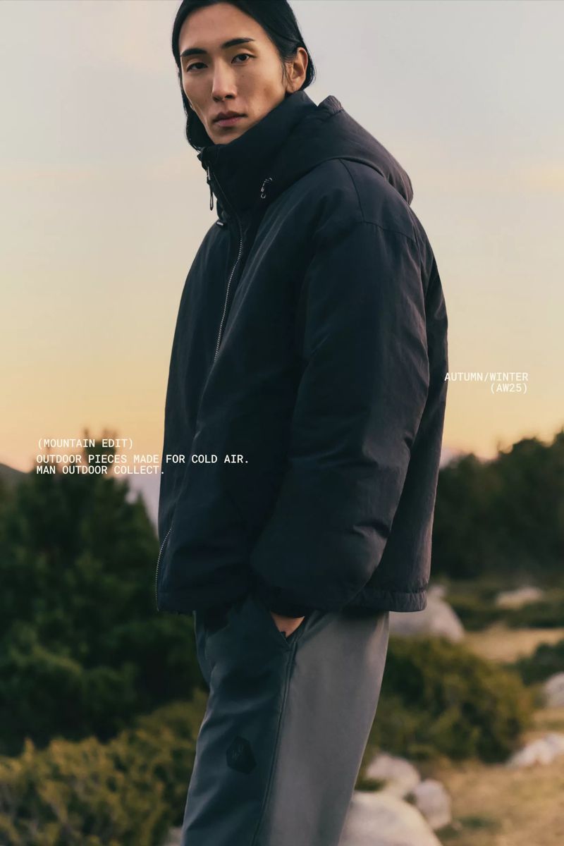 Asian man models a dark blue puffer jacket and grey trousers outdoors in a tan and green setting with soft, neutral lighting.