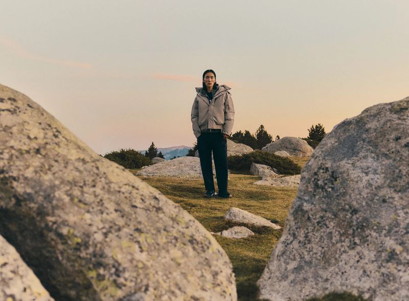 Adult male in a puffer jacket stands in a mountain field surrounded by large boulders, with trees and mountains in the background.
