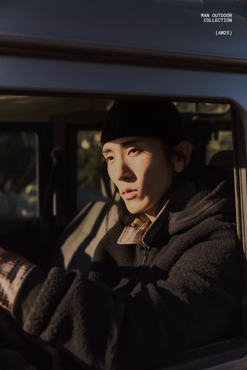 Adult Asian man in a Sherpa jacket and beanie sits inside a car with trees visible outside the car window in natural light.
