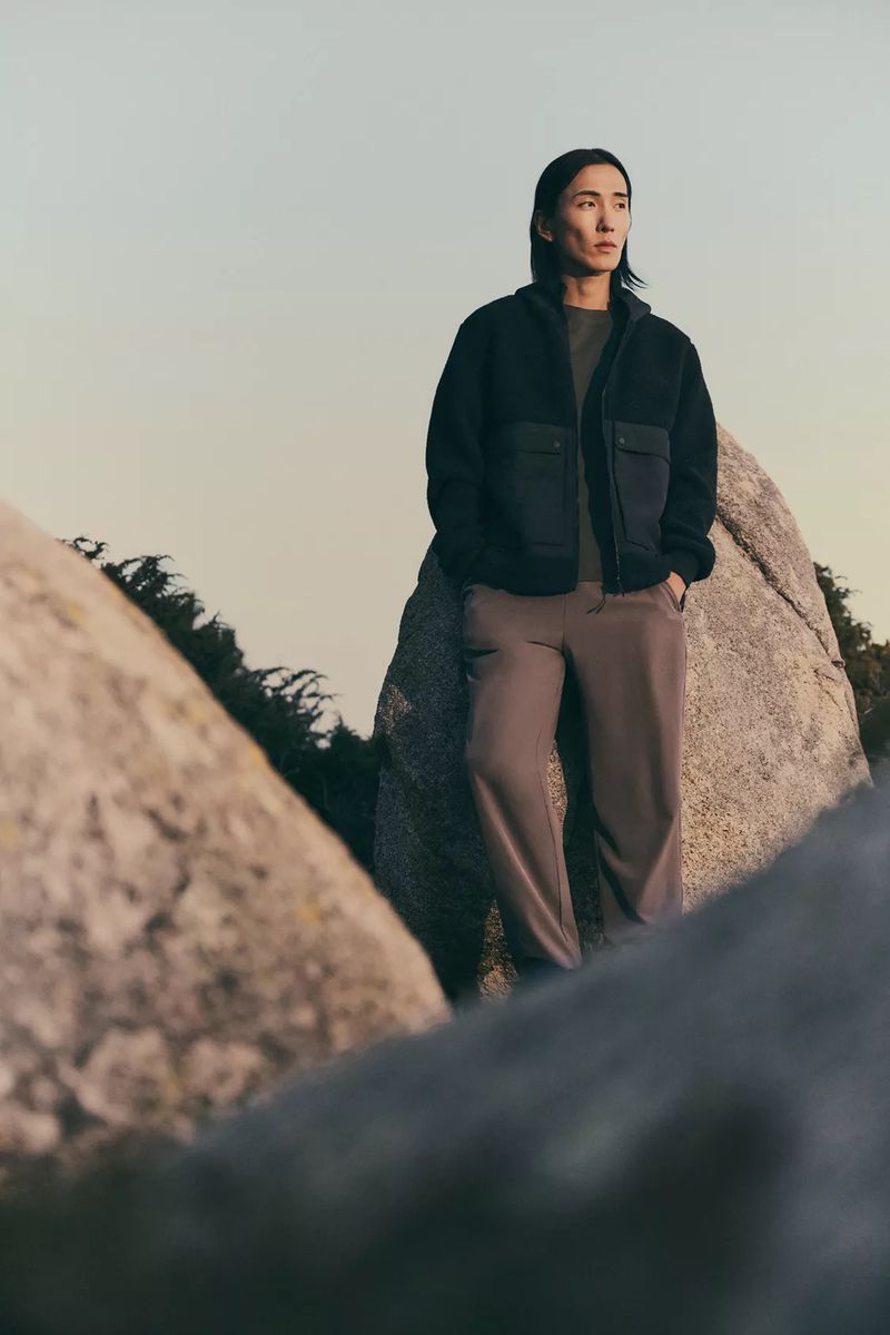 An adult male stands among large rocks wearing a black jacket, olive t-shirt, and grey-brown pants in an outdoor setting.