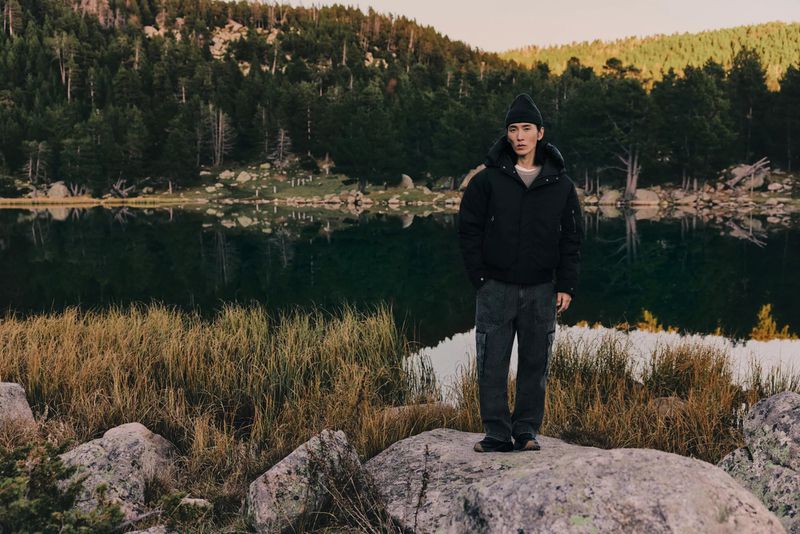 Man in a black jacket and beanie stands on rocks by a still lake, surrounded by a forest and tall grasses in a scenic outdoor setting.