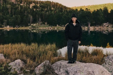 Model Hyuk Kim wearing a black hooded jacket and beanie standing on rocks in front of an alpine lake surrounded by pine forest.