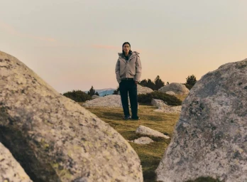 A model standing outdoors in a rocky meadow wearing a grey puffer jacket and dark trousers under an evening sky.