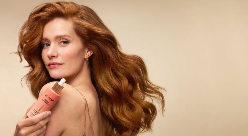 Red-haired woman poses indoors holding a Pantene hair product bottle in a beauty shot against a neutral backdrop.