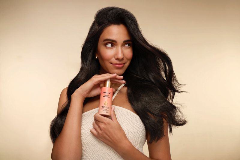 Brunette woman with long hair holds hair product. Beauty shot of a woman with a healthy hairstyle in front of a beige backdrop.