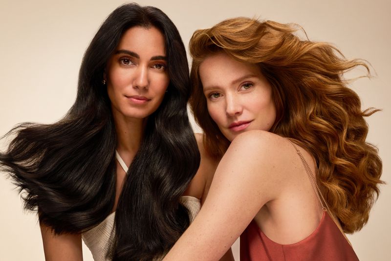 Two women pose indoors with flowing black and red hair against a beige backdrop, wearing casual tops and soft makeup for a beauty shot.