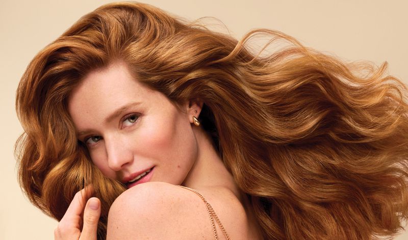 Close-up studio shot of a woman with red hair, gold earrings, and a delicate chain necklace. Her wavy hair is the focal point.
