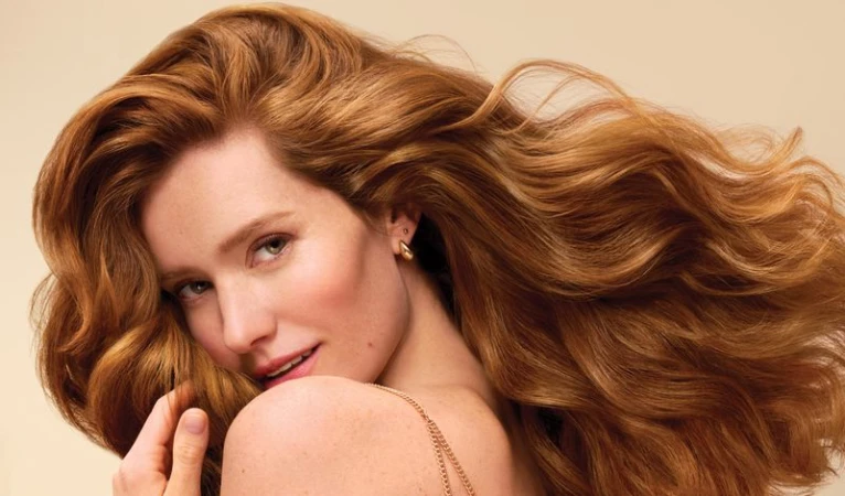 Portrait of a red-haired adult female with wavy long hair, light skin, and gold jewelry against a neutral background, beauty shot.