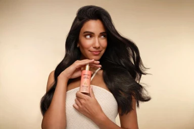 Woman with voluminous dark hair holding a Pantene Grow Abundant serum bottle against a warm beige background.