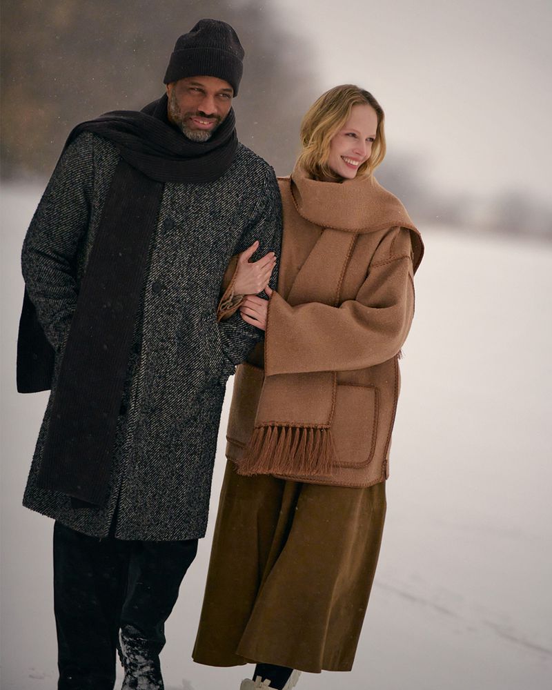A smiling couple walks arm-in-arm in a snow-covered landscape, wearing stylish coats and scarves in a light, overcast outdoor setting.