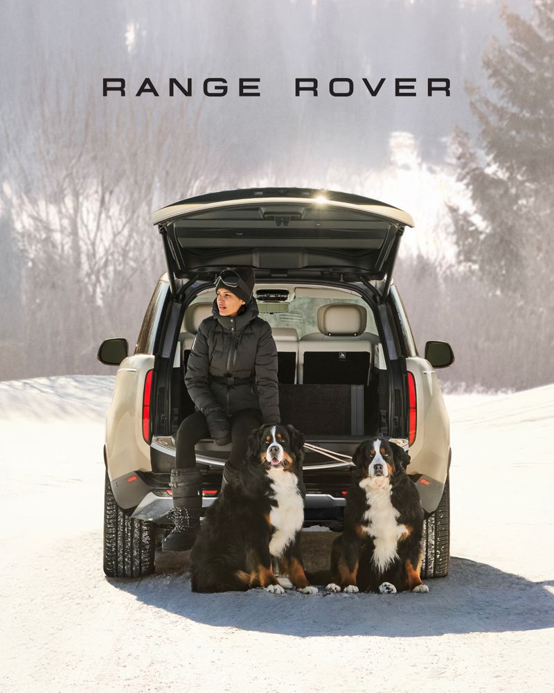 A woman and two Bernese Mountain Dogs pose in front of a Range Rover with its trunk open in a snowy, mountainous landscape.
